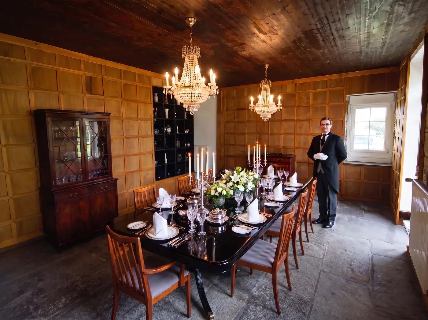 A formal dining room set for dinner with chandeliers, silver, crystal glassware, flowers and a butler in attendance
