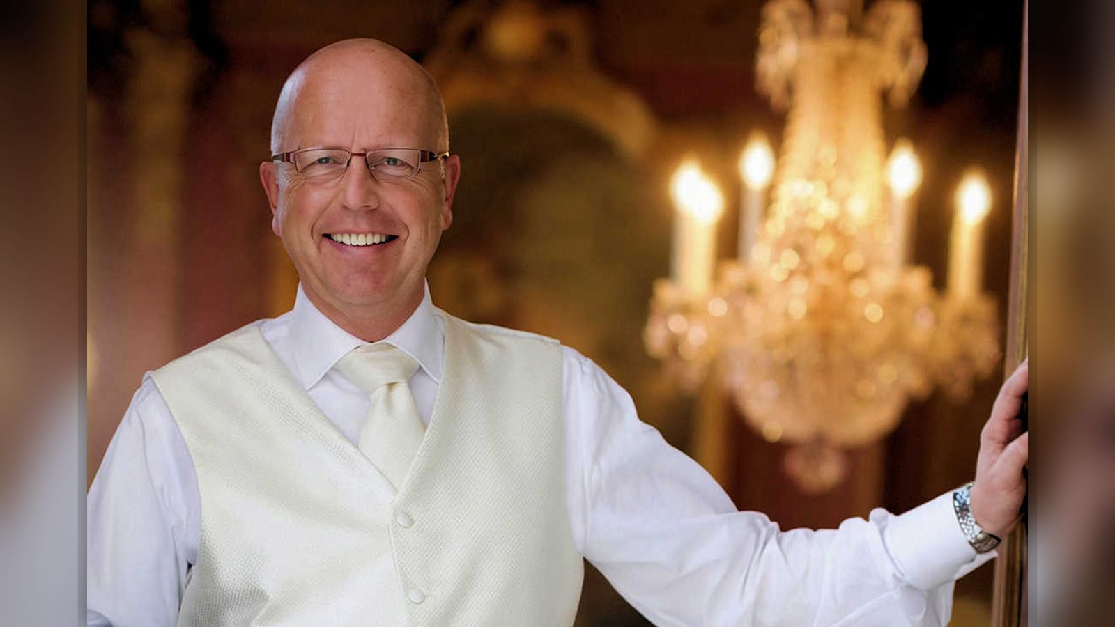 Robert Wennekes smiling, wearing clear-framed glasses, a white dress shirt and ivory waistcoat with cravat, photographed in a grand salon beside an ornate crystal chandelier.