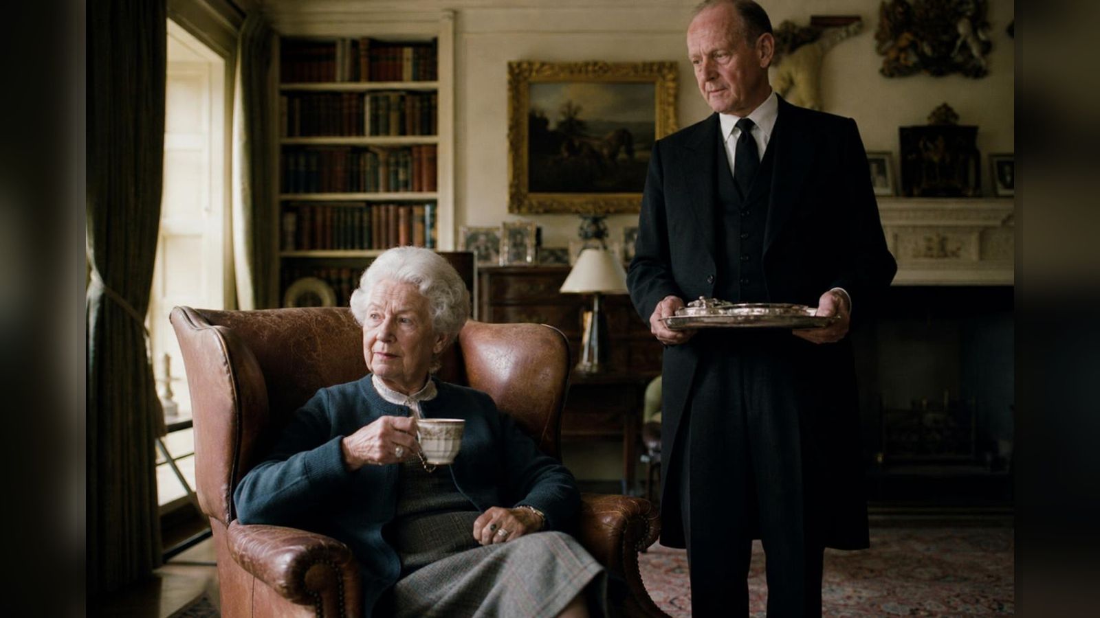 A long-serving butler in dark tails standing beside a silver tea tray, exchanging a warm smile with the elderly lady of the house seated by a fire in golden afternoon light.
